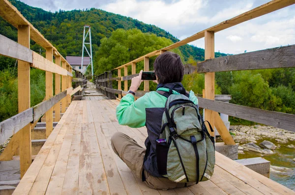 緑のジャケットとリュックを着た若者が山の近くの木製の橋の上に座り スマートフォンで写真を撮る — ストック写真