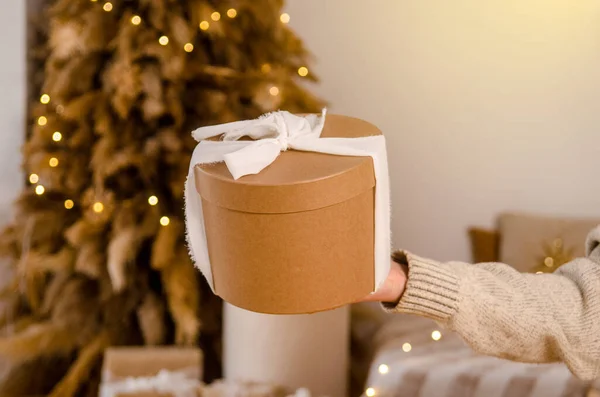Young European womans hand holding gift New Year cardboard round box with white fabric bow, with place for text or logo, in bright room with Christmas tree, bed, garlands
