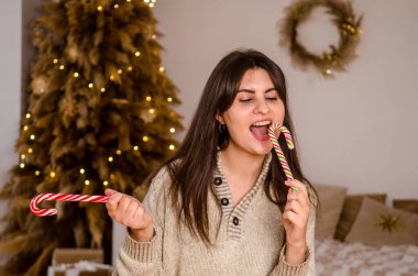 Süveter ve koyu renk saçlı genç bir kadın kırmızı beyaz bastonlarla oynar ve güler, parlak bir odada bir yatağın arka planına ve bir Noel ağacına karşı.