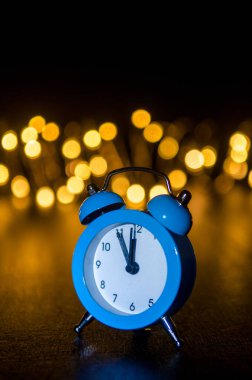Classic arrow blue alarm clock on a black background, illuminated from the side, behind round yellow blurred lights