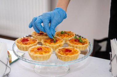 Peynirli, domatesli ve mikroyeşillikli tartlar, şeffaf bir stantta, bir etkinlikte yemek servisi, mavi eldivenli kadın eller mikroyeşillikler