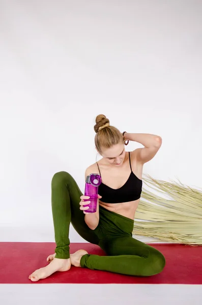 Tall slim European young woman with blond hair in a bun, no make-up, wearing a black top and green pants, doing sports or yoga, on a red carpet, palm leaf behind her, holding a purple bottle of water