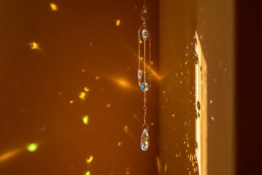 Metal altın zincirli cam güneş yakalayıcı, güneş ışığında parlıyor ve sarı arka planda gökkuşağını yansıtıyor.