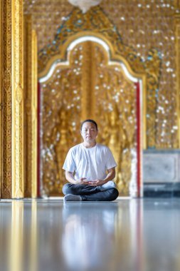 Asyalı adam, altın sarısı Tayland desenli bulanık arka planla tapınak kilisesine dökülen güneş ışığının yansımasıyla yerde meditasyon yapıyor..