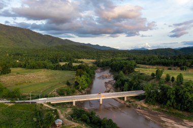 Nehir üzerindeki beton köprü bulutlu gökyüzünün altında orman nehri ve dağlarla çevrilidir..