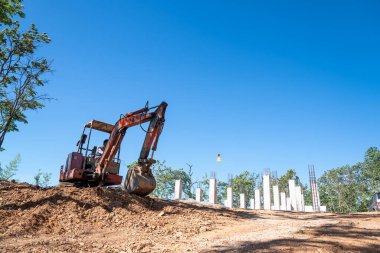 Hidrolik kazıcı, açık gökyüzünün altında bir sürü ağaçla çevrili bir inşaat alanında toprak kazıyor. Pençe kepçesi ağaç gölgesinin altında..