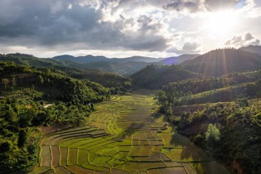 Tayland 'ın kuzeyinde, Omkoi bölgesinde, Chiang Mai bölgesinde, mavi bulutlu ve güneş ışığıyla yeşil dağların arasındaki teraslı tarım alanının havadan görünüşü.