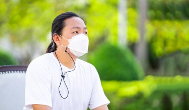 Beyaz tişört ve beyaz maske giyen bir adam koltukta sakince oturuyor. Kolları koltukta ve sırtı sırtüstü..