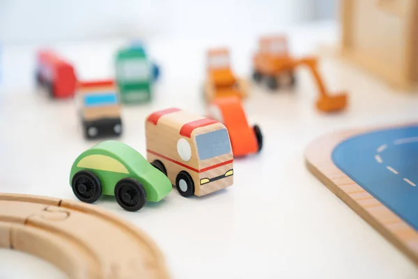 A Toy of wooden school bus drives beside a green car with many kinds of car behind it of a Vehicle Toy Set for children in an indoor playground.