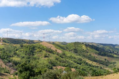 Güneşli bir günde, beyaz bulutlar ve mavi gökyüzünün altındaki çim tepesinin tepesindeki rüzgar türbinleri saf enerjiden elektrik üretiyordu..