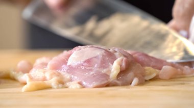 Mutfak bıçağı kullanan bir aşçı doğrama tahtasının üzerinde pişmemiş tavuk eti kesiyor, yemek malzemelerini hazırlarken yakından görülüyor..