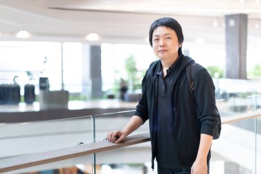 Standing next to the terrace barricade in a modern building, an Asian man exudes confidence in his stylish black attire. clipart