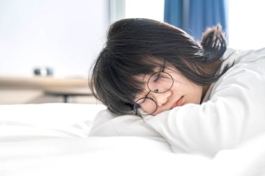 Asyalı genç bir kadın beyaz temiz bir yatakta beyaz tişörtle uzun kollu pijamalarıyla ve gözlerinde gözlük olmasına rağmen uyuyor..