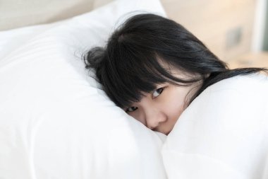 Beyaz uzun kollu beyaz pijamalı Asyalı hoş bir kadın yatağın kenarına uzanmış ve gözleri kameraya bakıyor.