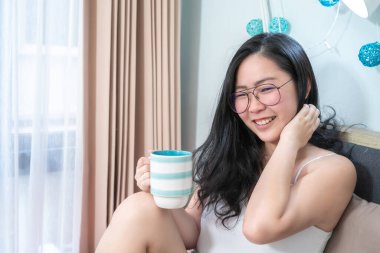 Asyalı Gözlüklü Kadın elinde bir fincan kahve tutuyor ve sabah yatak odasında dinleniyor..