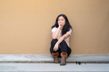 Relaxed Pretty Asian Woman sits on the rooftop floor with a light brown background and smiles to the camera. clipart