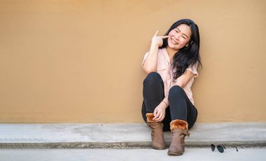 Rahat, güzel Asyalı kadın, açık kahverengi arka planıyla çatıda oturuyor ve kameraya gülümsüyor..