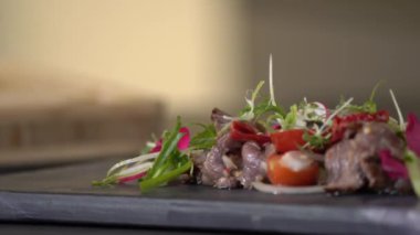 Baharatlı Tayland bifteği dilimlenmiş salatası çeşitli malzemeler kullanılarak hazırlanmıştır. Bunlar arasında yeşil, kırmızı turp ve pembe çiçekler de vardır. Dikdörtgen taş tabakta, düzenlenmiştir..