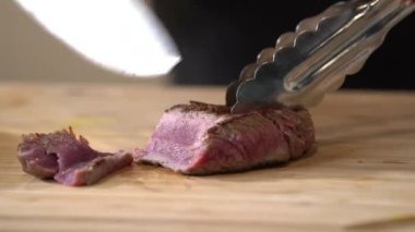 Orta pişmiş bifteği ahşap bir tahtanın üzerinde şef bıçaklarıyla kesen bir şefin yakın plan görüntüsü..