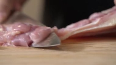 Mutfak bıçağı kullanan bir aşçı doğrama tahtasının üzerinde pişmemiş tavuk eti kesiyor, yemek malzemelerini hazırlarken yakından görülüyor..
