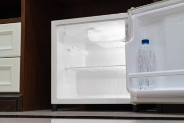 Küçük buzdolabı alçak açılı, boş rafların içinde ahşap tezgahın altında, ahşap dolabın yanında ve lüks bir otel odasında sandalye var..