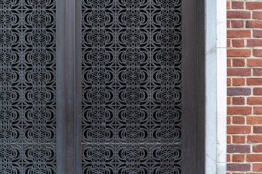 Siyah metal ızgara, geometrik desenli, kırmızı tuğladan bir duvara dayanıyor. Simetrik tasarım kalın bir mimari aksan ekler..