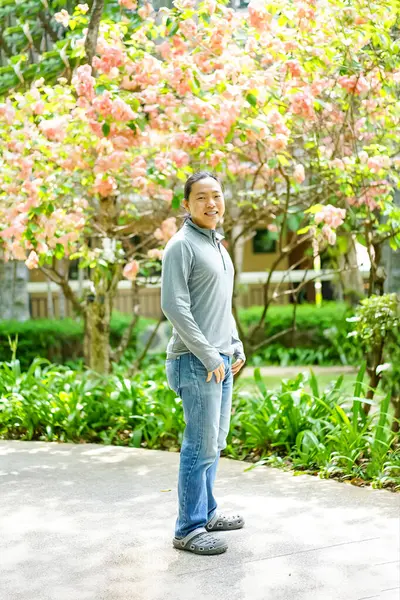 Asyalı uzun saçlı adam gri uzun kollu bir tişört giyiyor ve öğleden sonra ağaçta bir sürü çiçeğin önünde duruyor..