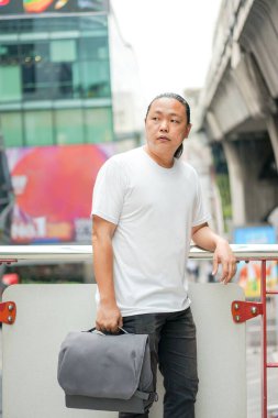 Uzun saçlı Taylandlı, Çinli bir adam teras barikatının önünde poz veriyor. Dışarıdaki bina ortamında sırtında gri bir leke var. 