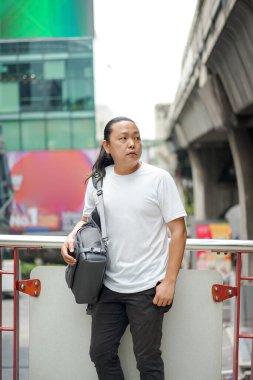 Uzun saçlı Taylandlı, Çinli bir adam teras barikatının önünde poz veriyor. Dışarıdaki bina ortamında sırtında gri bir leke var. 