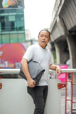 Uzun saçlı Taylandlı, Çinli bir adam teras barikatının önünde poz veriyor. Dışarıdaki bina ortamında sırtında gri bir leke var. 