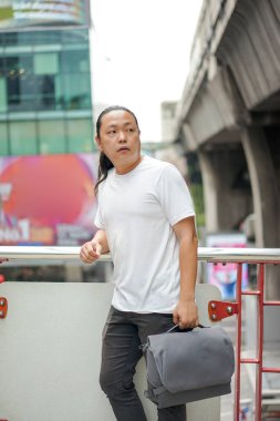 Uzun saçlı Taylandlı, Çinli bir adam teras barikatının önünde poz veriyor. Dışarıdaki bina ortamında sırtında gri bir leke var. 