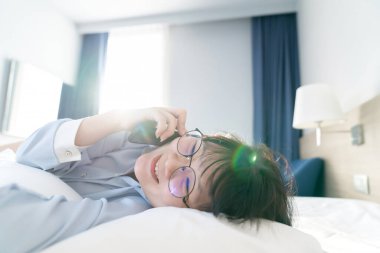 Asyalı genç gözlüklü kadın sabah yatağında yatarken cep telefonuyla konuşuyor..