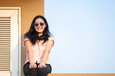 Güneş gözlüklü Asyalı kadın çatıdaki çimentoda oturuyor (tehlikeli değil çünkü zıt alan da çatıda, boş gökyüzünün önünde).