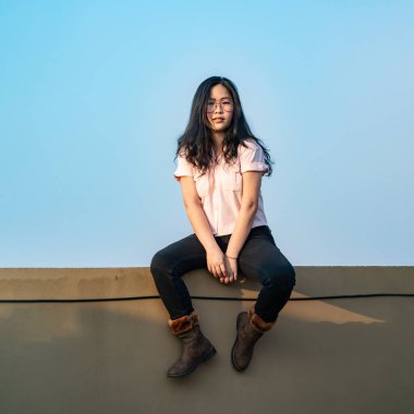 Gözlüklü Asyalı kadın çatıdaki çimentoda oturuyor (çatıdaki zıt alan da boş gökyüzünün önünde olduğu için tehlikeli değil).