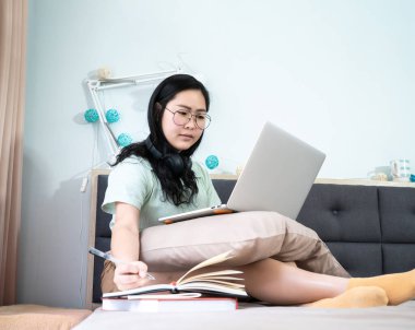 Gözlüklü güzel Asyalı kadın laptopuyla evden çalışıyor ve not defterine yanında pastel yeşili, kahverengi temalı bir not yazıyor..