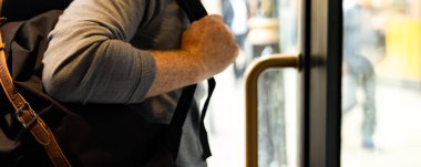 Kahverengi bir sırt çantası tutan, kapı kolunu tutarken otobüsten inen biri. Görüntü, şehir ortamındaki tipik bir banliyö anını yakalar