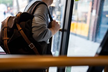 Kahverengi bir sırt çantası tutan, kapı kolunu tutarken otobüsten inen biri. Görüntü, şehir ortamındaki tipik bir banliyö anını yakalar