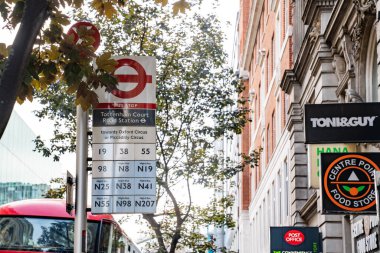 Londra, İngiltere, 12 Temmuz 2023, Tottenham Court Road İstasyonunda otobüs güzergahları ve gece otobüsleri listelenmiş, yakın dükkanlar ve arka planda kırmızı çift katlı bir otobüs