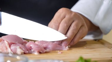 Mutfak bıçağı kullanan bir aşçı doğrama tahtasının üzerinde pişmemiş tavuk eti kesiyor, yemek malzemelerini hazırlarken yakından görülüyor..