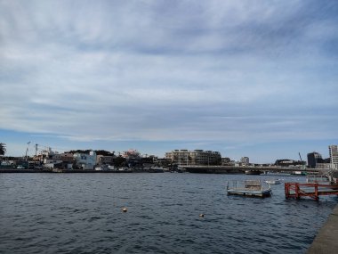 Kamakura, Japonya, 14 Mar 2024, Kıyı evleri ve yollarının yanında sahil boyunca yürüyen insanlarla kumsal. Sakin su, tekne ve bulutlu gökyüzü altındaki şehir binalarıyla liman manzarası.