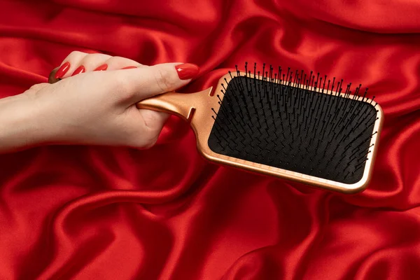 A red hairbrush in woman hand with red nails ion a red background. Beauty tools. Hair tools.