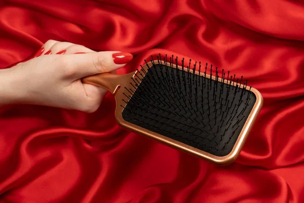 A red hairbrush in woman hand with red nails ion a red background. Beauty tools. Hair tools.