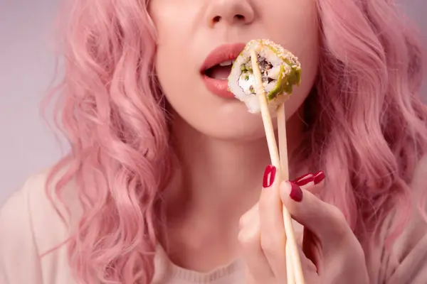 Young woman with red nails and pink hair is eating sushi close up.