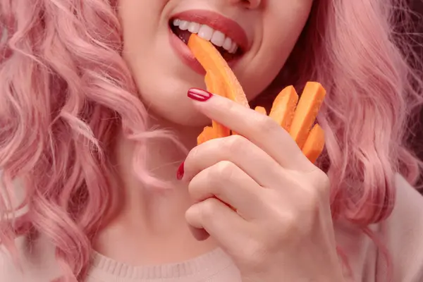 Carrots cut into strips in woman hand. Woman with pink curly hair is eating carrot close-up.