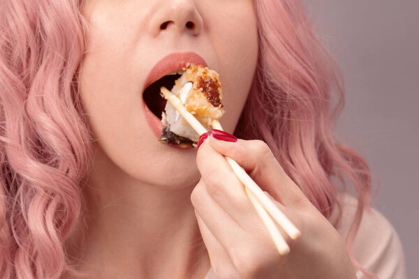 Молодая женщина с красными ногтями и розовыми волосами ест суши вблизи.