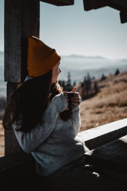 Genç bir kadın elinde bir fincan kahveyle dağların önünde oturuyor ve başka tarafa bakıyor.