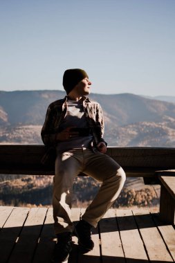 A young man in a beanie is sitting in a wooden house in front of a mountains landscape and looking away