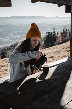 A young woman pouring out coffee from a Moka pot into a cup in outdoors house in front of a mountains landscape