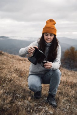 A woman in an orange beanie hat pouring out coffee from Moka pot into a cup outdoors in autumn time. Girl in a cozy casual sweater in front of a beautiful mountains view