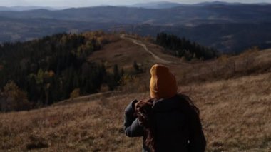 A woman in an orange beanie hat is standing in front of the mountains background. The girl is enjoying a beautiful sunny view of a road.